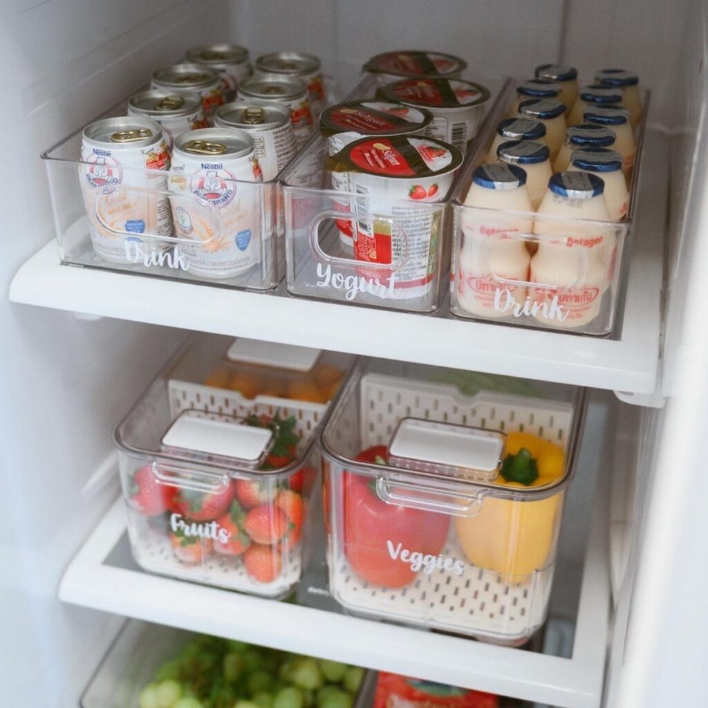 Clear plastic bins organizing drinks, yogurt, fruits, and vegetables inside a white refrigerator.