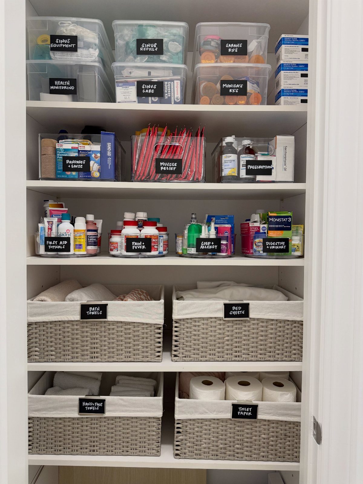 Organized white bathroom closet with labeled clear bins and woven storage baskets.