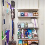 Organized cleaning closet with white shelves, clear labeled bins, and hanging vacuum and mops.