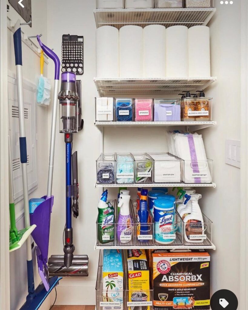 Organized cleaning closet with white shelves, clear labeled bins, and hanging vacuum and mops.