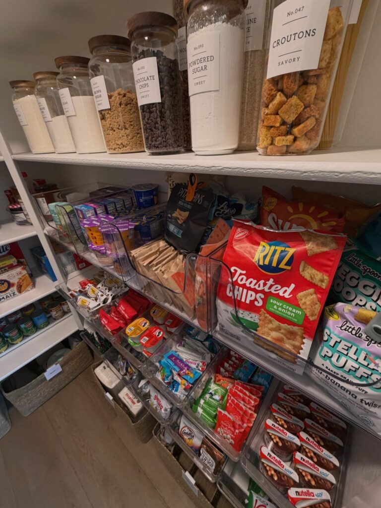 Organized pantry shelves with labeled glass jars and clear bins of various snacks.
