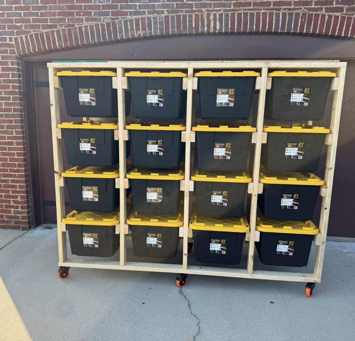Wooden rolling rack holding sixteen black storage totes with yellow lids outdoors.