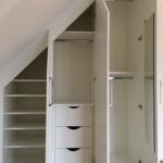 White built-in wardrobes under a sloped ceiling with shelving, drawers, and a full-length mirror.